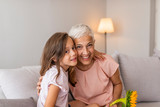 Granddaughter embracing her grandmother in living room. Lovely little girl with her grandmother looking at the camera. Girl and grandma on a sofa Smiling senior woman and girl embracing