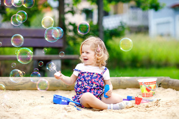 Beautiful little blonde toddler girl having fun with blowing soap bubble blower. Cute adorable baby...
