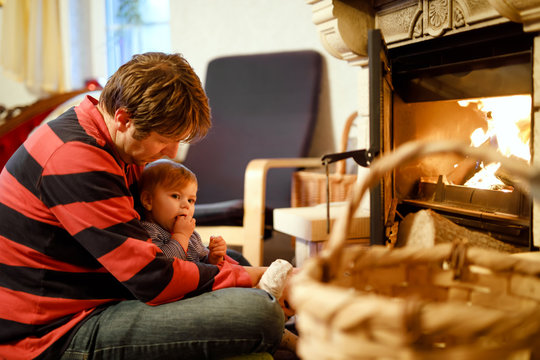 Middle age father sitting by fireplace with cute little baby girl at home. Happy family, dad with daughter make fire for Christmas time. Safe handling for kids - Powered by Adobe