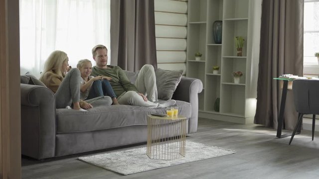 Tracking left shot of beautiful family of three laughing and talking while watching tv series sitting on sofa at home. Mother, father and little daughter enjoying spending leisure time together