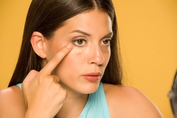 Fototapeta premium Young woman applying concealer on her low eyelids on yellow background