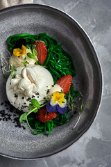 Burrata salad with strawberries and seaweed, on a gray background. Salad with buratta cheese on a gray plate and concrete marble background.