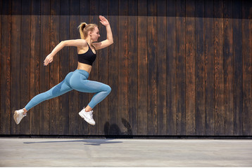 Young sporty beautiful woman with the right body, jumping and running, hurry on a wooden wall background. Gymnast jumping high with a toothy smile, full length, on the street