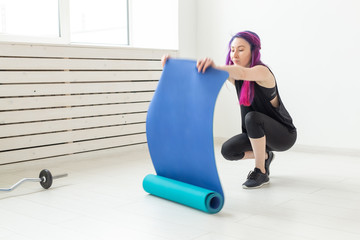 Slim young girl hipster with colored hair lay a gymnastic mat on the floor In the bright gym before the start of training. Concept of a slim healthy body and sports lifestyle.