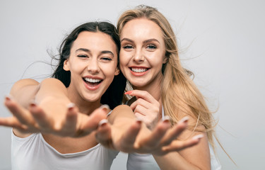 Joyful cute girlfriends in white tank tops smiling and stretching hands to camera isolated white...