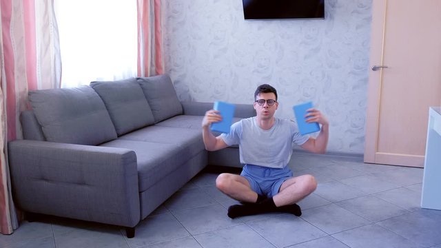 Funny nerd man in glasses is doing exercises for hand biceps with yoga blocks instead of dumbbells sitting on the floor at home. Sport humor concept.