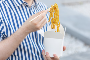 An unidentified young male hipster with a mustache and beard is eating chinese noodles with wooden chopsticks from a white lunch box. Asian cuisine concept.