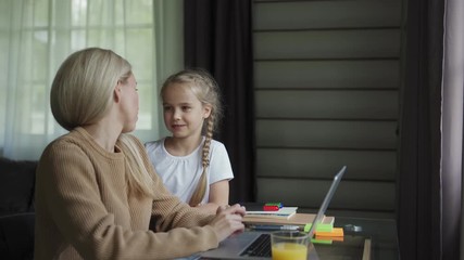 Tracking right shot of cute little daughter coming to loving mother sitting at desk and working on laptop at home. Mother and daughter talking and embracing warmly - Powered by Adobe