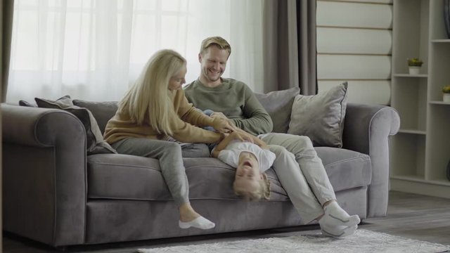 Happy family of three spending leisure time together sitting on sofa at home. Mother and father tickling playful little daughter laughing cheerfully