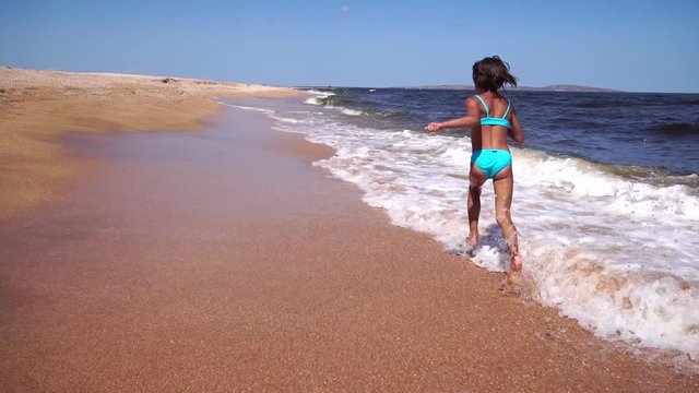 Happy little girl runs along the beach on the water on the sea shore. Kid is running on the beach, raises splashes