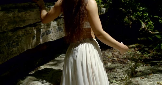 slender woman with dark long hair is strolling in woodland in summer, back view