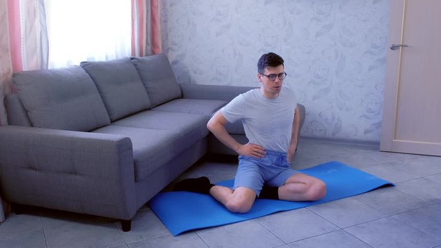 Funny nerd man in glasses is doing torso twists exercise sitting on mat at home. Sport humor concept.