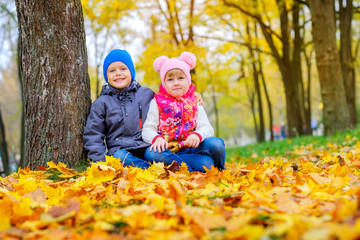 Cute children in a beautiful autumn park are sitting on foliage near a tree.