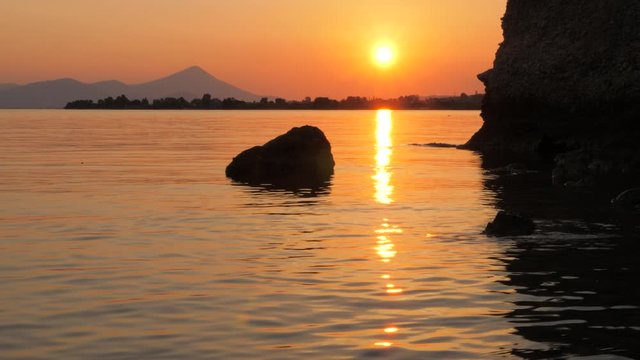 Seascape and rockes with sunset on background. Sea water reflecting sunset with mountains on background. Sunset on sea horizon with stons and mountains. The sun goes down Time lapse