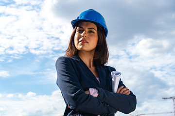 Low angle portrait of a beautiful and successful lady architect wearing a blue safety helmet and posing outdoors with crossed arms