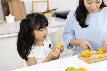 Obraz na płótnie Canvas Happy Asian family Little girl eating Orange fruit and Mother are preparing the vegetables and fruit in the kitchen at home. Healthy food concept