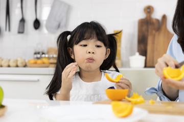 Happy Asian family Close up of Little girl eating Orange fruit and Mother are preparing the vegetables and fruit in the kitchen at home. Healthy food concept