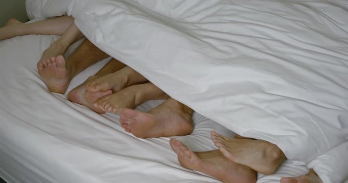 two men and three women are lying in bed together, moving legs under blanket, close-up