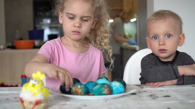 Cute little siblings look at painted Easter eggs while mother work in kitchen