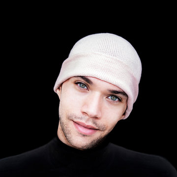 Portrait of a handsome smiling young man with green eyes wearing a pale pink woolen cap, black background