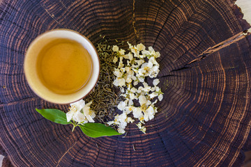 Obraz na płótnie Canvas Cup of tea, green tea, dried blossoms and sprig of jasmine on wooden board. Flat lay with space for text