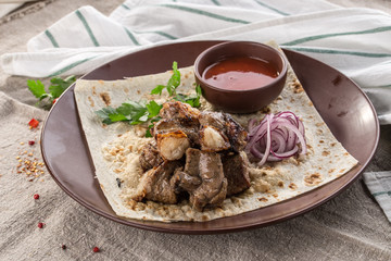 Shish kebab on lavash bread with red onion and tomato sauce on brown plate on tablecloth