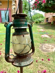 Fototapeta na wymiar Old lantern, broken and rusty oil lamps hanging on pole, background of green grass, illuminated by summer evening sun light beams