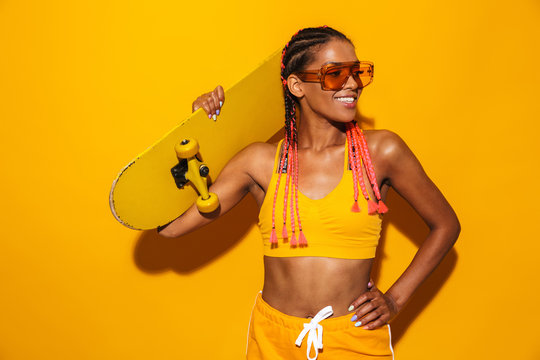 Image of nice african american woman wearing sunglasses holding skateboard and smiling
