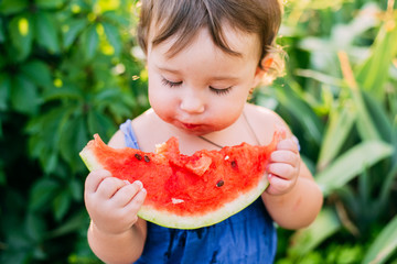 Charming little girl eating watermelon in the yard on the background of plants