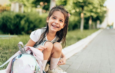 Outdoors portrait of happy little girl preschooler smiling broadly and sitting on doorstep with backpack after school. Happy child pupil relaxing after kindergarten. People and education concept