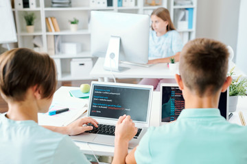 Two schoolboys and schoolgirl sitting by desks in front of computers