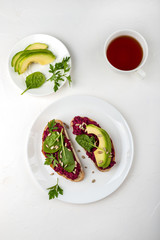 Breakfast. Bruschettas with beetroot hummus, avocado, spinach and seeds on a white plate and cup of tea. Top view.