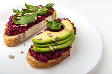 Bruschettas with beetroot hummus, avocado, spinach and seeds on a white plate on a white background.
