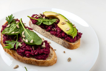 Bruschettas with beetroot hummus, avocado, spinach and seeds on a white plate on a white background.