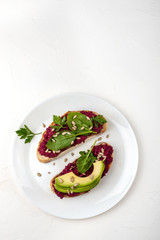 Bruschettas with beetroot hummus, avocado, spinach and seeds on a white plate on a white background.