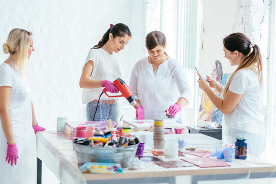 Woman painter holding hair dryer while female beginnres studying creating fluid acrylic abstract painting in art therapy class, dropping paints on canvas.
