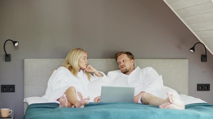 Married middle aged couple in white bathrobes lying on bed in hotel room, relaxing and talking. Man using laptop computer - Powered by Adobe