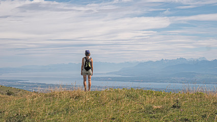 A lone female hiker stands on a mountain top peacefully looking at the surrounding views of an alpine lake, valley and high mountain peaks.