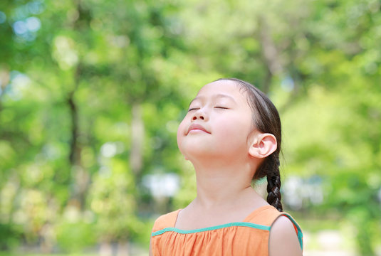 Portrait of happy Asian child close their eyes in garden with Breathe fresh air from nature. Close up kid girl relax in green park for good health.