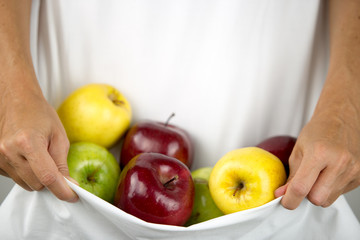 A Caucasian woman holds some apples of various types and colors in her white dress clasped in her hands