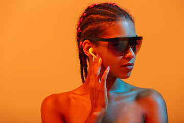 Pretty beautiful young african naked woman posing isolated over yellow neon lights wall background listening music with headphones wearing sunglasses.