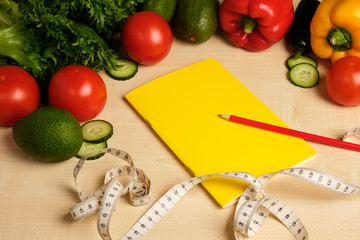 Green and red vegetables lie at the top of the frame above a yellow notepad, red pencil and measuring tape on a wooden table.
