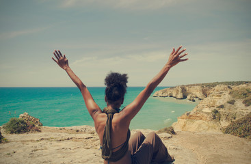 Black woman with open arms looking at the sea sitting on a cliff in Algarve, Portugal