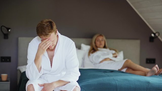 Rack focus of upset woman in white bathrobe lying on bed looking displeased. Depressed husband or lover rubbing forehead in foreground and looking at camera