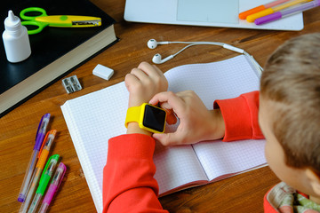 Little boy sitting at the table and looking smart watch. Smart watch for baby safety. The child makes school lessons, listening to music, calling friends.