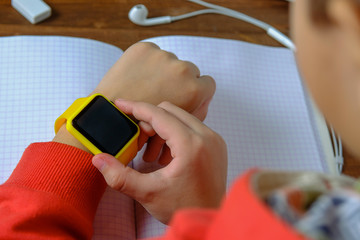Little boy sitting at the table and looking smart watch. Smart watch for baby safety. The child makes school lessons, listening to music, calling friends.