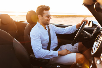 Photo of attractive caucasian man driving convertible car by seaside at sunrise - obrazy, fototapety, plakaty