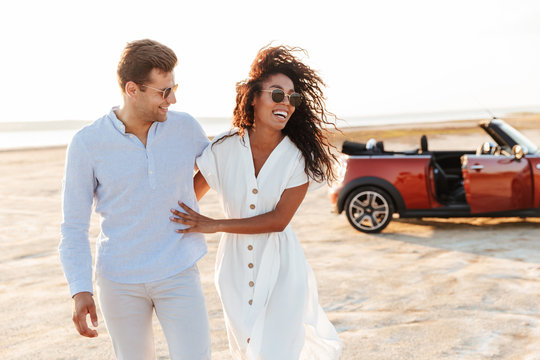 Photo of adorable multiethnic couple smiling together while walking by car on beach