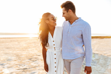 Photo of elegant multiethnic couple smiling and hugging together while walking on beach by seaside