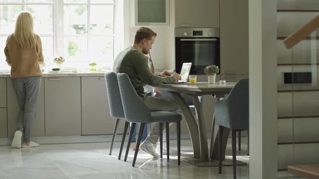 Tracking left shot of middle aged man using laptop computer sitting at table in domestic kitchen. Little daughter running downstairs to him, caring mother giving her glass of orange juice and kissing